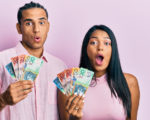 Young latin couple holding australian dollars scared and amazed with open mouth for surprise, disbel
